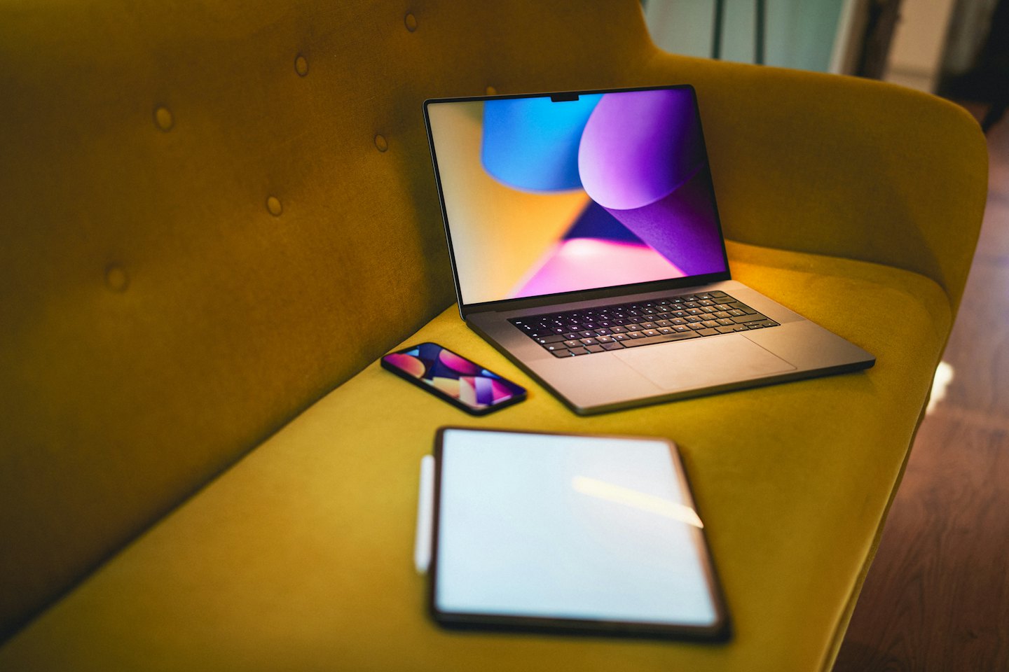 Silver laptop, smartphone, and tablet on a yellow sofa.